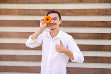 Blossoms, Emotions, Gesture And People Concept - A Young Caucasian Guy In A White Shirt Showing Thumbs Up And Holding An Orange Flower Instead Of An Eye On A Brown Ribbed Background With Copy Space
