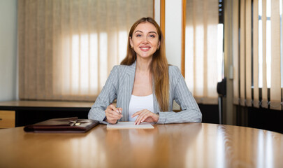 Wall Mural - Female lawyer sitting at desk and looking in camera while filling out information in document.