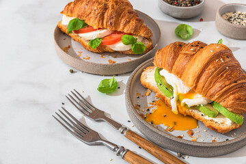 Wall Mural - Tasty breakfast with croissant sandwiches with poached egg, avocado, soft cheese, mozzarella and tomato in a plate