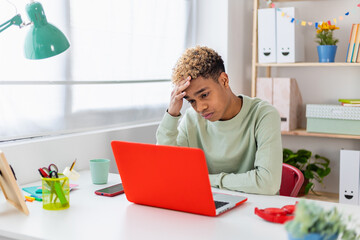 Portrait of stressed teenager boy sitting on desk in youth room - Young worried male student reading bad news on laptop - Education concept