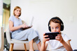 © khmelev - Child watching cartoons on the phone while the mother is working on the laptop. Schoolboy holds mobile phone in his hands. Distance learning. Cybersport.