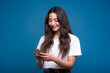 A young and attractive caucasian or arab brunette girl in a white t-shirt using a mobile phone isolated on a blue studio background.