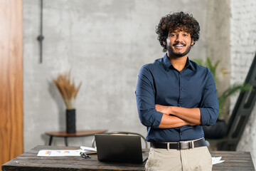 Smiling curly middle eastern businessman standing near desk and looking at the camera. Young positive male student with modern flat on background. Proud and successful mixed-race small business owner