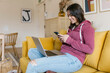 © Westend61 - Smiling young woman sitting with laptop text messaging through smart phone on sofa
