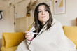 © Westend61 - Beautiful woman wrapped in blanket holding thermometer in living room