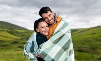 Poster - love, travel and tourism concept - happy smiling couple in warm blanket over farmland fields and hills at wild atlantic way in ireland background