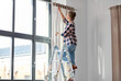home improvement, decoration and renovation concept - happy smiling woman on ladder hanging curtains