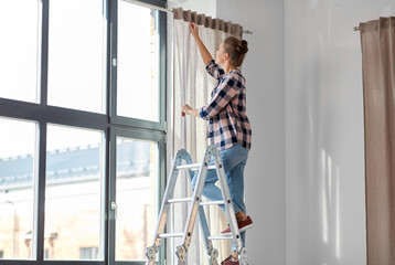 home improvement, decoration and renovation concept - happy smiling woman on ladder hanging curtains