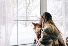 Young Caucasian Happy Woman Sitting Near Window With Her Dog In Winter Holidays, Looking Outside.