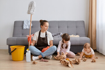 Attractive dark haired woman wearing white t shirt, brown apron and jeans, sitting on floor near cough with her children making mess, female holding mop in hands, doing household chores.