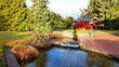 malerischer Teich mit Brücke und roter Terrasse mit Gartenhaus und hohen Fichten in traumhafter Natur