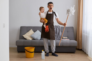 Wall Mural - Full length portrait of man wearing casual attire and brown apron cleaning house with baby and screaming, looking at camera with shocked aggressive face, working about the house with kid.