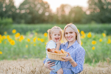 Wall Mural - Adorable mother and daughter in romantic dresses eat fresh pastries in a wheat field. Summer family portrait. Eco.