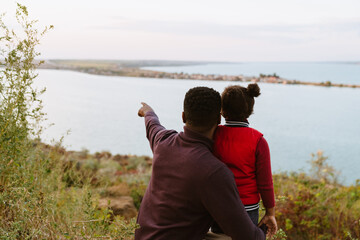 Wall Mural - Black man pointing finger away while looking at river with his daughter