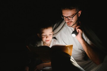 Happy family reading bedtime story under blanket in evening. Father and son spend time together. Father's Day.