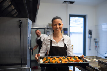 Happy female cook looking at camera and holding tray with baked pumpkin pieces in commercial kitchen