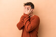 © Asier - Young caucasian man isolated on beige background blink through fingers frightened and nervous.