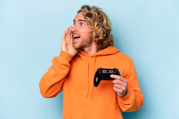 Wall Mural - Young caucasian man playing with a video game controller isolated on blue background shouting and holding palm near opened mouth.