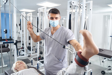 Wall Mural - Man medical worker supporting pensioner leg during exercise