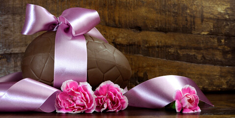 Wall Mural - Happy Easter chocolate easter egg with satin pink ribbon and pink flowers against a dark vintage recycled wood timber background with copy space.e. Sized to fit popular social media and web banner.