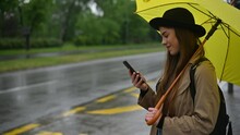Woman Using A Smartphone And Holding A Yellow Umbrella On A Bus Stop On A Rainy Day