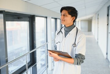 Wall Mural - Portrait of confident young Arabian Indian male medical doctor in white coat, standing with clipboard in hands on background of modern hospital building outdoor