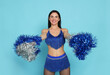 Beautiful cheerleader in costume holding pom poms on light blue background