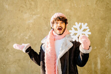 Happy Hispanic Man With Excite Surprise Expression In Pink Winter Knitted Hat, Scarf And Gloves Showing Artificial Snowflake Looking At Camera Standing On Green Background. Winter Season Sale Concept.