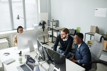 Two young program developers in casual clothes sitting in armchairs by desk in front of computer monitors and working over new program