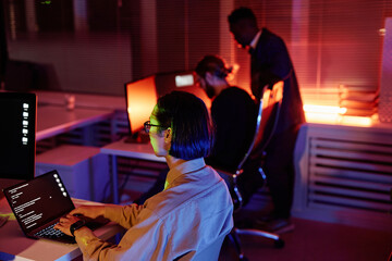 Young Asian software developer working over new program while sitting by desk and looking at laptop screen against two colleagues