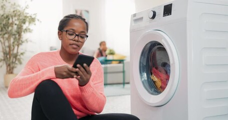 Wall Mural - Multifunctional washing machine controlled by mobile app, fast program, woman closes drum with colorful clothes, taps fingers on phone, walks away and sits on couch next to daughter