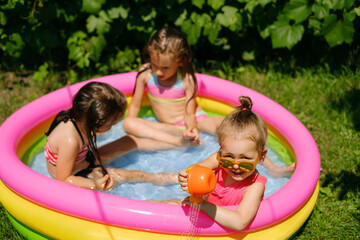 Three girls are having fun in a small inflatable swimming pool in the garden on a hot sunny day. Children play with a watering can in the water. Outdoor recreation