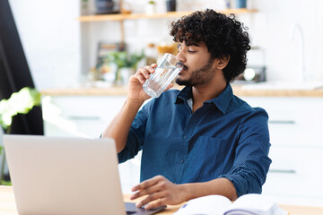 Wall Mural - Young Indian man drinking still mineral water. Male freelancer working, using a laptop, drinks clean water. Healthy lifestyle concept