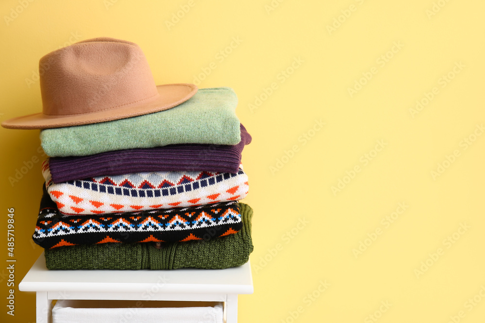Stack of stylish sweaters and felt hat on stool near color wall