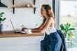 © undrey - Serious woman sitting at table and typing on laptop in kitchen
