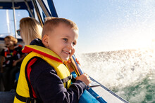 Portrait Of Cite Little Blond Happy Excited Smiling Caucasian Boy Wear Lifevest Enjoy Sailing On Motor Boat Sea Against Blue Sky And Water Splash Wave Sun Backlit. Summer Travel Vacation Recreation