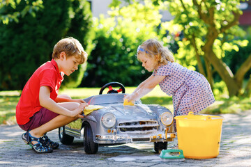 Wall Mural - Two happy children washing big old toy car in summer garden, outdoors. Brother boy and little sister toddler girl cleaning car with soap and water, having fun with splashing and playing with sponge.