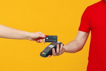 Wall Mural - Cropped delivery guy employee man wear red T-shirt uniform work as dealer courier hold wireless bank payment terminal customer process acquire credit card payment isolated on plain yellow background.