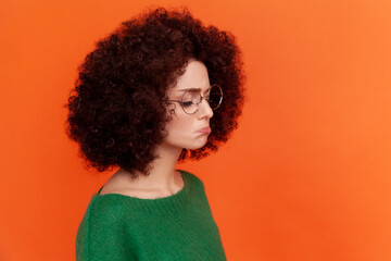 Wall Mural - Profile portrait of sad upset woman with Afro hairstyle wearing green casual style sweater standing with pout lips, feels sadness, cry. Indoor studio shot isolated on orange background.