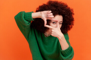 Young adult woman with Afro hairstyle wearing green casual style sweater standing crop composition focus hand gesture and looking at camera. Indoor studio shot isolated on orange background.