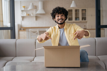 Young smiling Indian man unpacking box with present sitting at home. Online shopping, delivery service concept. Successful blogger influencer recording video pointing fingers on carton box, copy space