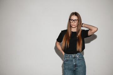 Pretty young woman with vintage glasses in trendy clothes with fashion black t-shirt and stylish jeans poses near a white wall