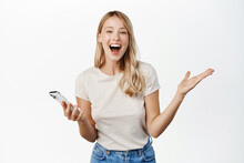 Happy Blond Girl Winning On Mobile Phone App, Watching Smartphone And Reacting With Joy And Amazement To Notification, Standing Over White Background
