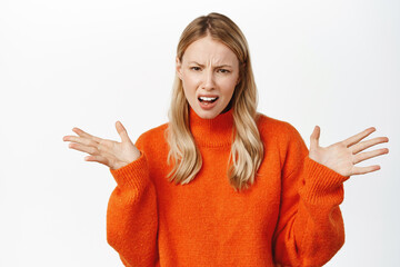 Wall Mural - Confused and annoyed young blond woman, staring puzzled at camera, standing in red sweater over white background. Whats your problem
