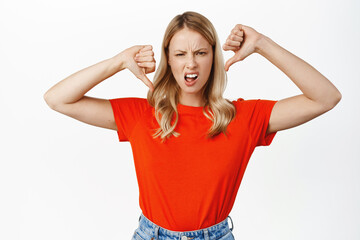 Wall Mural - Disappointed blond girl shows thumbs down, dislike, disapproval sign, negative feedback, standing in red tshirt over white background
