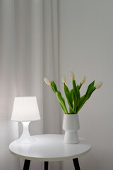 Wall Mural - White tulips in a white vase on a table with a table lamp