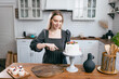 © primipil - Pastry chef confectioner young caucasian woman in gray dress with knife cut slice cake on kitchen table. Cakes cupcakes and sweet dessert Scandinavian style studio