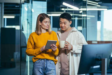 Wall Mural - Two employees are having fun. during the break an Asian man and a blonde woman, colleagues work together in a modern office, look at a tablet computer together