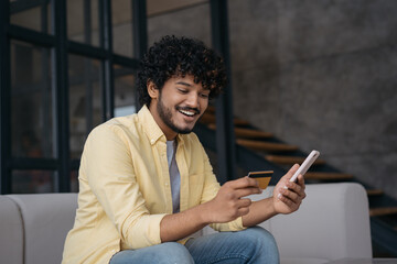 Smiling Indian man holding credit card using mobile phone shopping online, check card balance. Mobile banking. Emotional asian guy ordering food online looking at digital screen sitting at home 