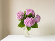 © Natalie - Close up of pink hydrangea flowers in glass vase on white table against neutral wall background (selective focus)